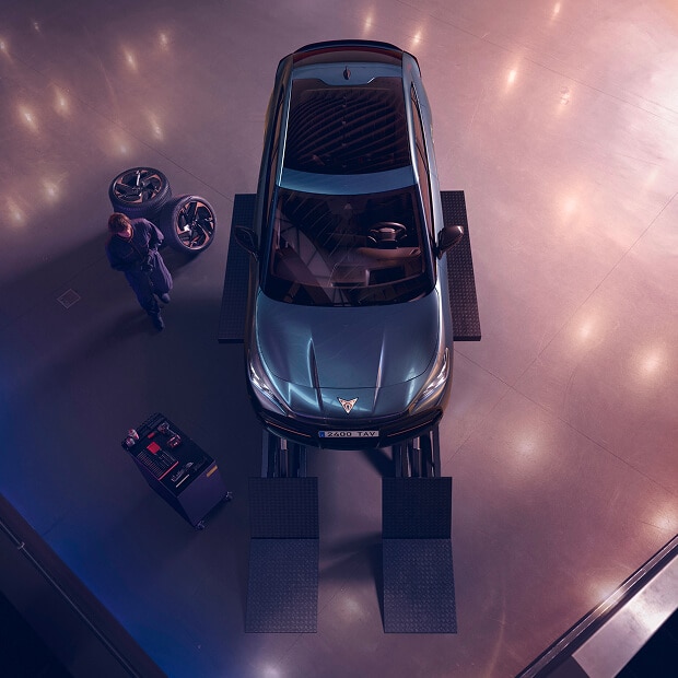 An overhead view of a CUPRA vehicle on a lift in a service garage. A person is visible near some car wheels on the left, and a tool cart is near the front of the car. The floor has subtle light reflections.